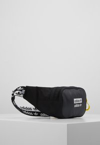 Cinturón negro de Adidas con parche de logo en el frente, bolsillo con cremallera y correa ajustable con marca sobre una superficie blanca contra un fondo liso.