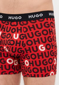 Boxer-briefs rouges arborant un motif répétitif de logo "HUGO" en noir et blanc, avec une taille noire embossée de "HUGO" en blanc.