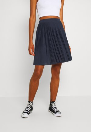 Personne portant un débardeur court blanc, une jupe plissée bleu marine longueur genou et des baskets montantes noires, debout devant un fond uni.