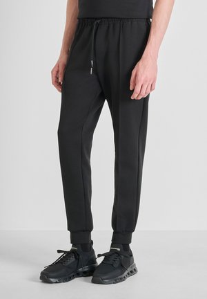Schwarze sportliche Jogginghosen aus glattem Material, mit einem elastischen Bund und Kordelzug sowie gerippten Bündchen. Getragen mit schwarzen Sneakern.