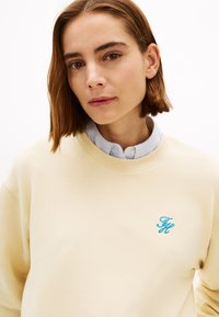 Φούτερ σε κρεμ απόχρωση με υφή, που διαθέτει ένα μπλε κεντημένο λογότυπο στο στήθος. Φορεμένο πάνω από ένα ανοιχτό μπλε πουκάμισο με γιακά.