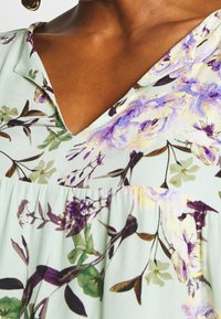 Sukienka w kwiaty z dekoltem w kształcie litery V w jasnym miętowym kolorze. Ozdobiona wielokolorowymi kwiatami w odcieniach purpury i żółci, z gromadzonym fasonem w talii.