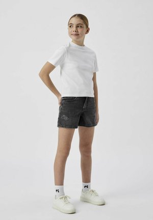 T-shirt blanc à manches courtes, short en denim noir avec des détails usés, et baskets blanches avec des chaussettes à motifs. Tenue simple et décontractée.