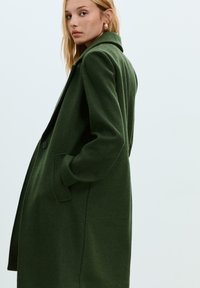Zelený vlněný kabát s vystouplým límcem, zapínáním na jeden knoflík, bočními kapsami a strukturovaným střihem, vystavený na světlém pozadí.