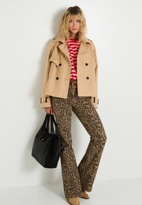 Femme portant un trench-coat court beige, une chemise rayée rouge et rose, un pantalon à imprimé léopard, des bottes beiges, tenant un sac à main noir contre un mur blanc.