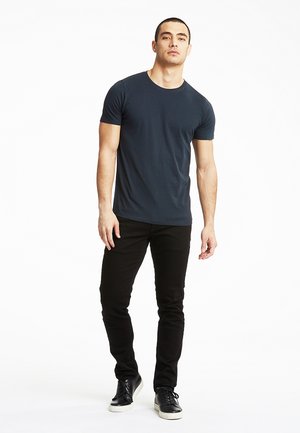 Mężczyzna stojący, ubranego w granatowy t-shirt z krótkim rękawem, czarne spodnie i czarne tenisówki z białymi podeszwami na białym tle.