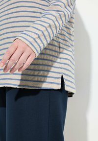 Langärmliges Hemd in Beige mit blauen Streifen, mit abgerundetem Saum und seitlichen Schlitzen, kombiniert mit dunkelblauen Hosen.