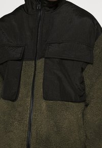 Fleecjas met een zwart bovenstuk en een olijfgroen onderstuk. Inclusief twee voorste klepzakken en een rits sluiting.