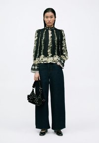 Schwarze Bluse mit Blumenmuster und langen Ärmeln, mit hohem Kragen und gemusterten Bündchen, kombiniert mit dunklen, weit geschnittenen Jeans und einer kleinen schwarzen Handtasche.