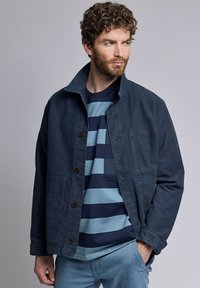 Veste en velours côtelé bleu marine avec col, fermeture à boutons sur le devant, deux poches latérales, portée par-dessus une chemise rayée bleu marine et bleu clair.