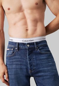 Blugi din denim de culoare albastru închis cu un brâu alb Calvin Klein care prezintă textul logo-ului negru. Design clasic cu cinci buzunare și o uzură subtilă.