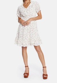 Blommönstrad klänning i vitt, med korta ärmar med knytband, smockad midja och en volangkant. Kombinerad med bruna platåklackar.