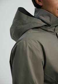 Szara wodoodporna kurtka z wysokim kołnierzem i kapturem, wykonana z gładkiego, wytrzymałego materiału o minimalistycznym wzornictwie i szwach.