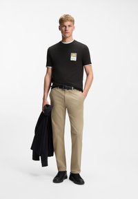 Νεαρός άνδρας που φοράει μαύρο μπλουζάκι με σχέδιο, μπεζ παντελόνι, μαύρα αθλητικά παπούτσια, κρατώντας ένα μαύρο μπουφάν στο ένα χέρι, στέκεται μπροστά σε λευκό φόντο.