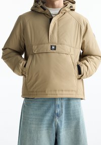 Beige geïsoleerd jack met een capuchon, ritssluiting, voorzak en elastische manchetten. Gedragen over lichtblauwe spijkershorts.