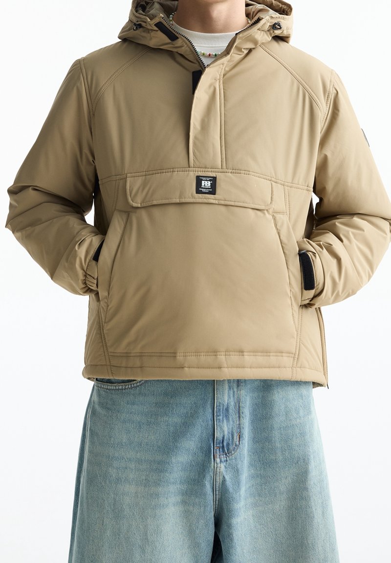 Beige geïsoleerd jack met een capuchon, ritssluiting, voorzak en elastische manchetten. Gedragen over lichtblauwe spijkershorts.