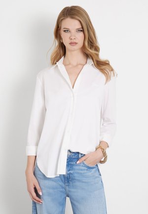 Camisa blanca de manga larga con botones, de tela suave, con un corte ajustado y un cuello sutil, combinada con jeans de mezclilla azules, que tienen un estilo relajado.