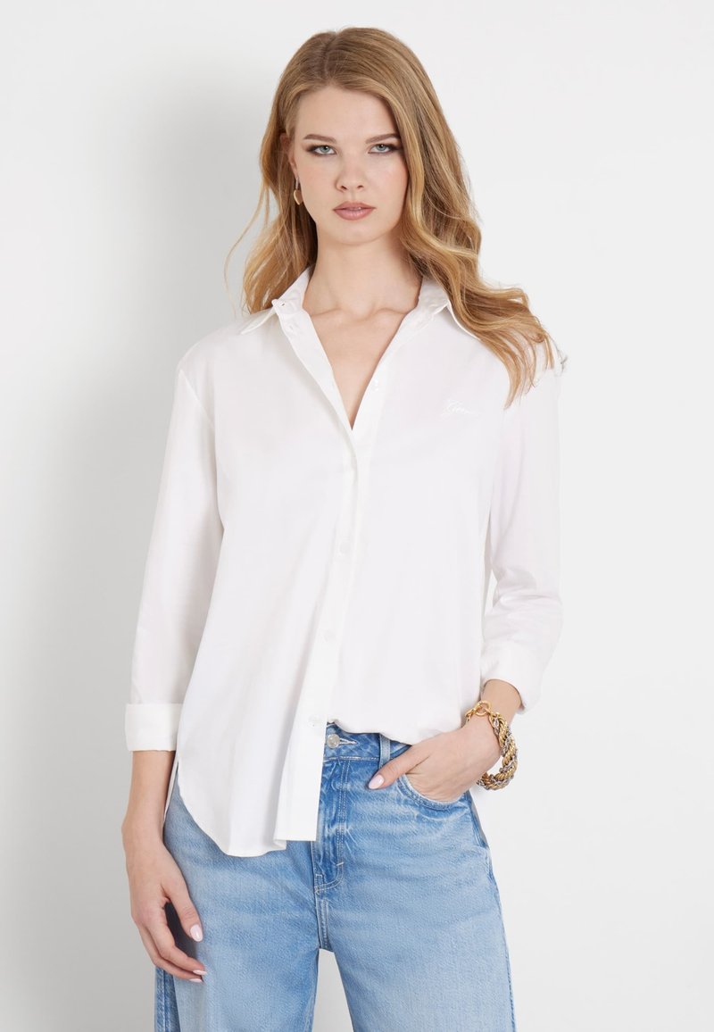 Camisa blanca de manga larga con botones, de tela suave, con un corte ajustado y un cuello sutil, combinada con jeans de mezclilla azules, que tienen un estilo relajado.