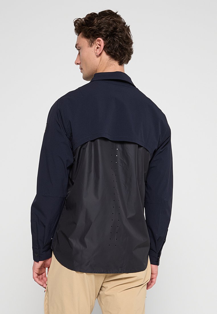 Dunkelblaue Jacke aus leichtem, strukturiertem Stoff. Verfügt über ein kontrastierendes Rückenteil mit Belüftungslöchern. Lange Ärmel.