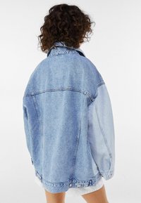 Velká denimová bunda ve světle modré barvě s kontrastními odstíny, špičatým límcem a knoflíkovými uzávěry na manžetách a lemu. Textura je hladká a ležérní.