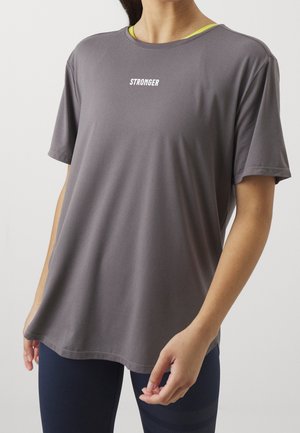 Personne portant un t-shirt ample gris à manches courtes avec "STRONGER" imprimé sur la poitrine et des leggings bleu marine, debout devant un fond uni.