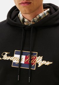 Sudadera negra con logo bordado "Tommy Hilfiger" en dorado, que presenta un diseño rectangular en rojo, blanco y azul. Detalles de cordón visibles.