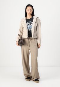 Veste en velours côtelé beige avec capuche sur un t-shirt graphique noir, associée à un pantalon large beige et des baskets noires à semelles crème. Sac à main marron.