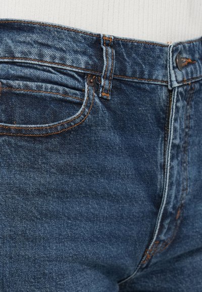 Közelkép kék farmernadrág derék részéről, narancssárga varrással, övbemélyedésekkel, elülső zsebbel és gombos záródással, fehér bordázott felső ellenében.