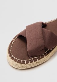 Sandale din material textil maro cu curele încrucișate, talpă din jute împletit și detalii cusute pe margine. Textură moale și design plat.