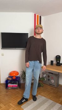 Μακρυμάνικο καφέ πουκάμισο, ανοιχτό μπλε τζιν, μαύρα αθλητικά παπούτσια. Εσωτερικός χώρος με πορτοκαλί λάμπα και διακοσμητικά στοιχεία στο παρασκήνιο.