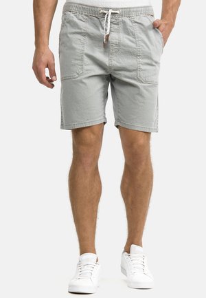 Mann trägt hellgraue Shorts mit Kordelzug und Fronttaschen sowie weiße Turnschuhe, steht mit Händen in der Nähe der Taschen.