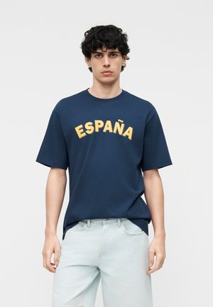 Νέος άνδρας με μαλλιά σγουρά που φοράει μπλε σκούρο T-shirt με τη λέξη "ESPAÑA" σε κίτρινο και ανοιχτό μπλε τζιν, μπροστά από απλό φόντο.