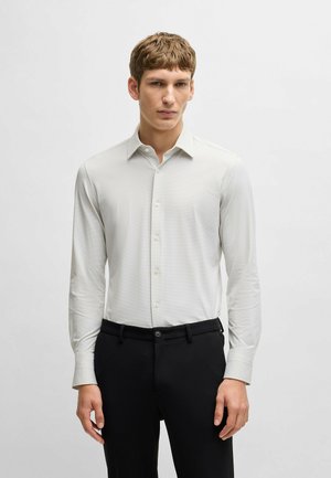 Camicia bianca a maniche lunghe con un motivo sottile, colletto a punta e chiusura con bottoni, abbinata a pantaloni neri e una cintura di tessuto.