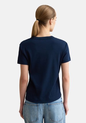 Mujer con coleta rubia, vistiendo una camiseta azul marino lisa de manga corta y vaqueros de mezclilla azul claro, vista desde atrás.