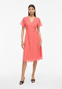 Krátke rukávy, zavinovacia šaty v koralovej farbe s textúrou chevron. Má výstrih do V, bočný viazací pás a dĺžku po kolená. Štylizované s čiernymi opätkami.