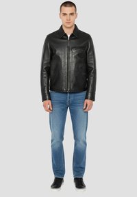 Veste en cuir noir avec fermeture éclair, col pointu et poches latérales. Portée avec un jean bleu clair et des baskets noires.