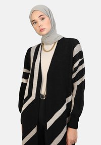 Long black cardigan with beige diagonal stripes, made of knitted fabric. Worn over a light beige top, featuring a gold chain necklace.
