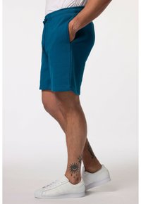 Tyrkysové šortky s elastickým pasem a šňůrkou, mají boční kapsy. V kombinaci s bílými sportovními botami a viditelnými tetováními na nohou.