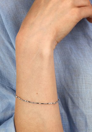 Silbernes Armband mit einem schlanken, gewundenen Design, das das Licht reflektiert. Am Handgelenk getragen, vor einem hellblauen, strukturierten Stoffhintergrund.