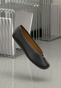 Ballerina nera in pelle con una texture liscia, punta a mandorla e dettaglio del logo dorato di lato, esposta in un contesto minimalista.