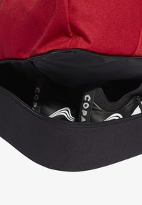 Bolsa de tela roja con un compartimento con cremallera negra que contiene zapatos negros con acentos blancos y un logo. Textura suave, forma estructurada.