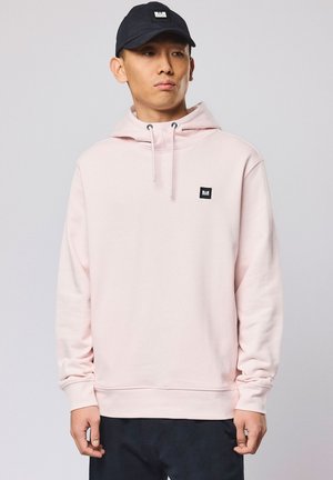 Junger Mann trägt einen hellrosa Hoodie mit Kordeln und einem kleinen schwarzen Logopatch, kombiniert mit einer schwarzen Mütze und dunkler Hose, steht still.