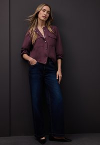 Chemise bordeaux boutonnée avec deux poches poitrine, associée à un jean large bleu foncé et des ballerines noires, sur un fond sombre.