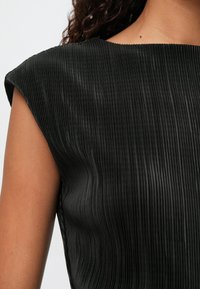 Μαύρη πλισέ μπλούζα με φαρδιά λαιμόκοψη και μανίκια τύπου καπάκι, που αναδεικνύει ένα υφασμάτινο σχέδιο με υφή και ραφές που δημιουργούν διακριτές κάθετες γραμμές.