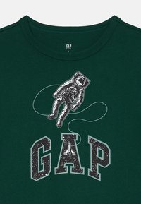 Grünes Kurzarm-T-Shirt mit Rundhalsausschnitt, das ein Astronautenmotiv und darunter das Wort "GAP" in fetten, gesprenkelten Buchstaben zeigt.