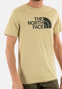 Beige bavlněné tričko s velkým černým logem "The North Face" na přední straně. Kulatý výstřih a krátké rukávy s hladkou texturou.