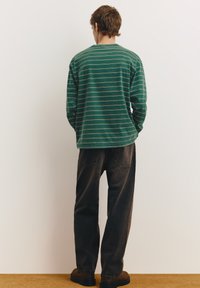 Cămașă verde cu mâneci lungi, cu dungi subțiri bej, asortată cu blugi închiși la culoare, largi și cizme maro. Văzută din spate.