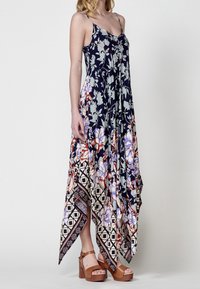 Robe longue avec un fond bleu marine orné de motifs floraux dans des tons plus clairs. Ourlet asymétrique avec une bordure à motif géométrique. Bretelles fines.