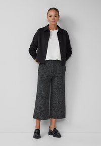 Schwarze Cropped-Jacke, weißes Hemd und weit geschnittene gemusterte Hose in Schwarz und Grau. Glänzende schwarze Loafers. Minimalistisches Design und klare Linien.