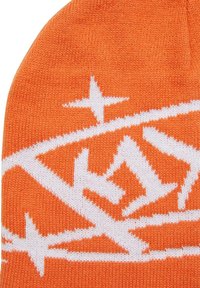 Narancssárga kötött sapka fehér geometrikus mintázattal és csillag díszítéssel. Puha anyagból készült, textúrált felülettel.